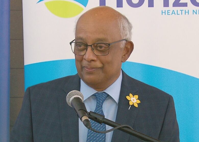 A man wearing a suit with a yellow daffodil pin. There is a microphone in front of him. 