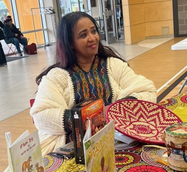 A woman wearing traditional African attire sits at a table with books and a drum and other items.