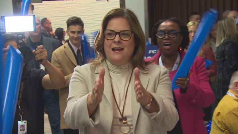 Woman in white suit, with happy supporters in the background, brings her hands together to clap.