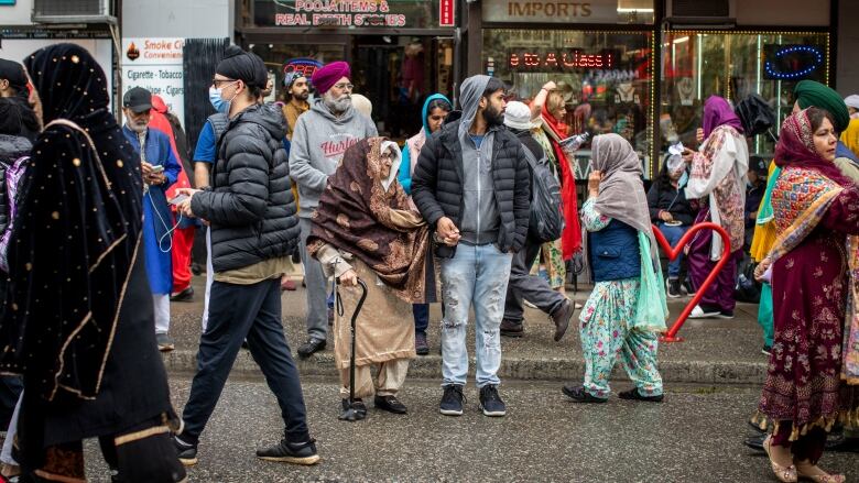 People line up along the street with Indian stores visible behind them.