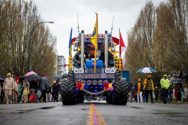 A large tractor is festooned with gold flags and festive attachments at the head of a parade of people.