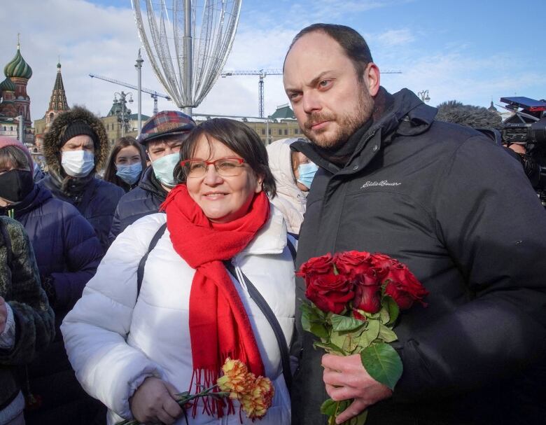 A woman and a man each holding flowers are shown posing in an outdoor photo.