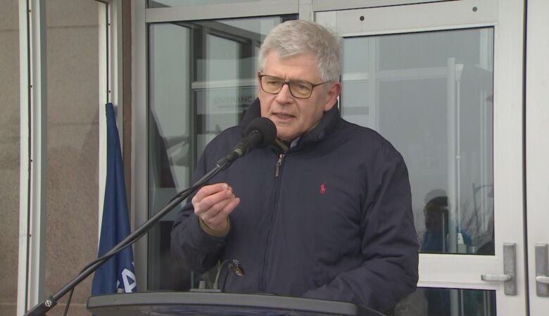 A grey-haired man in a dark jacket speaks at a lectern outside glass doors.