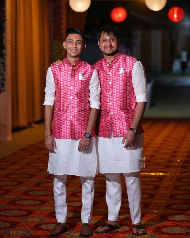 Two men pose for a photo together. Both men are wearing red patterned shirts and long white tunics and white pants underneath. 