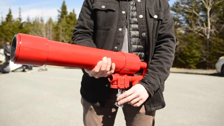 A man wearing a black jacket and brown pants holds a red plastic tube with a trigger on the bottom. He is standing on pavement in front of trees.