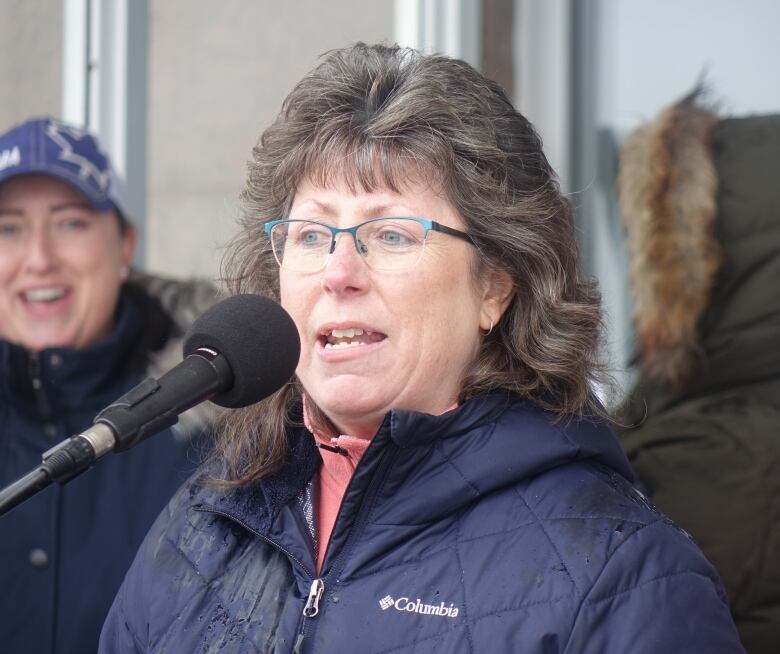 A woman in glasses and a blue coat speaks into a microphone.