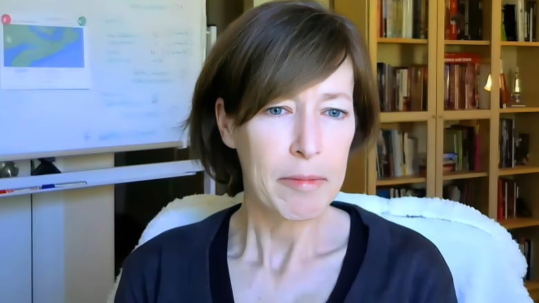 A woman is shown speaking to the camera with a bookcase behind her.