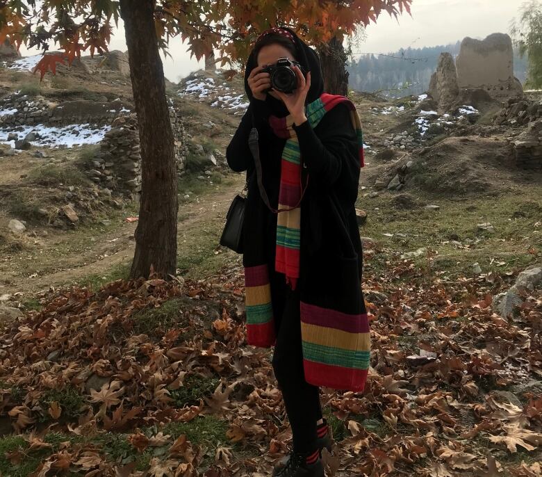 A woman with dark hair stands outside in a field of leaves with a camera to her eye.