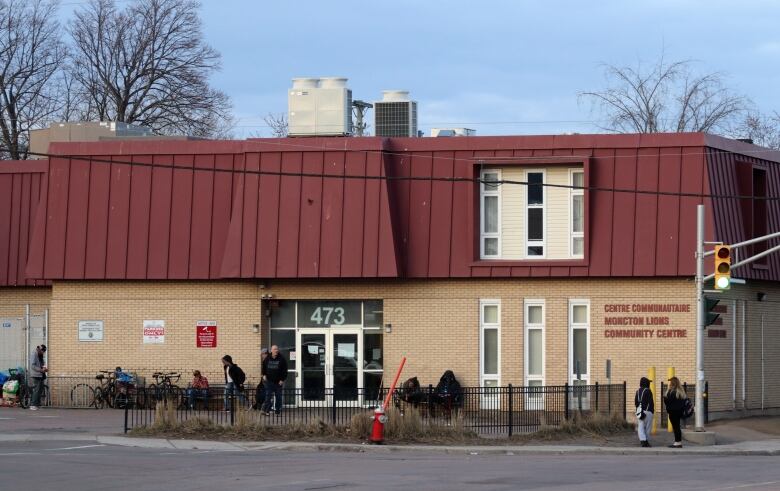 Several people standing outside a two-storey building made of yellow bricks a red metal roof.