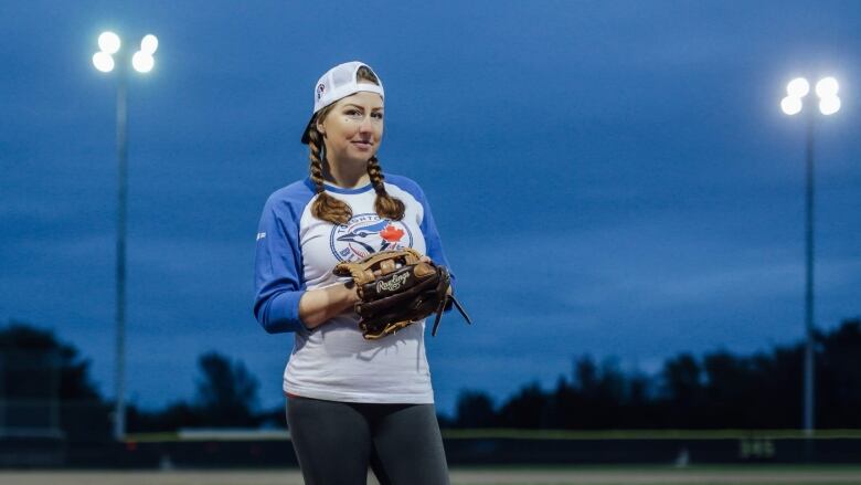 A woman wearing a Toronto Blue Jays shirt, a baseball cap and baseball glove stands in a baseball field under bright stadium lights.