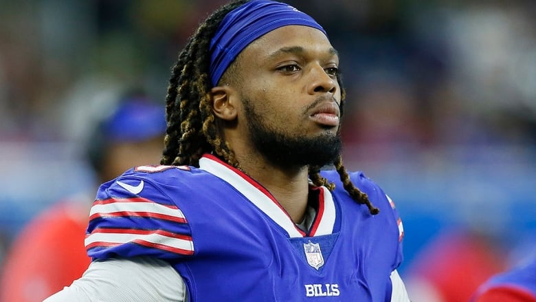 Men's pro football player, sporting a blue headband, looks onto the field from the sideline.