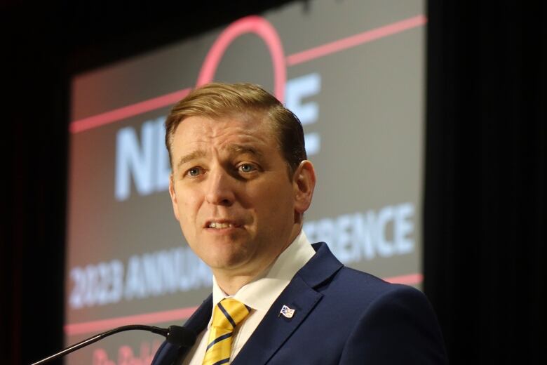 A man in a suit, stands in front of a small microphone, against a backlit screen.