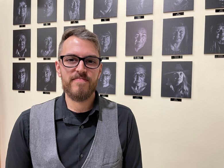 Wayne MacDonald stand in a sweater vest and glasses smiling in front of a wall of portraits he captured 