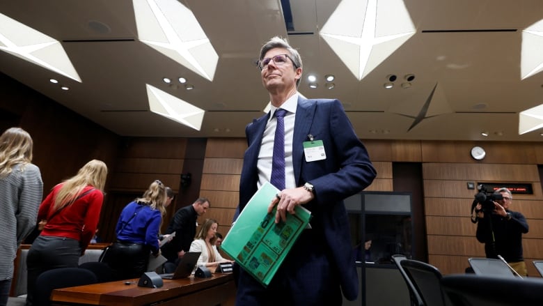 A man in a suit stands in a conference room, holding some papers. 