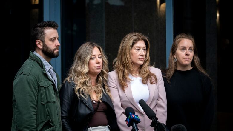 Four people with solemn expressions stand together outside a glass window on an overcast day.