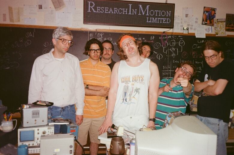 Seven men pose in a grainy photo. Behind them is a blackboard with mathematical formulas on it. Above is a sign that says 