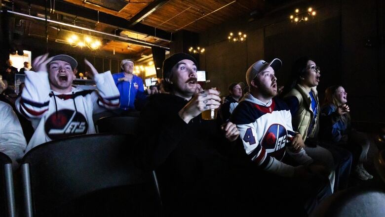 Winnipeg Jets fans, some wearing jerseys, exclaim with open mouths while sitting in theatre seats and watching a hockey game on a big screen.