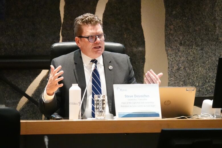 A man in a suit gestures while sitting in front of a laptop and a microphone.