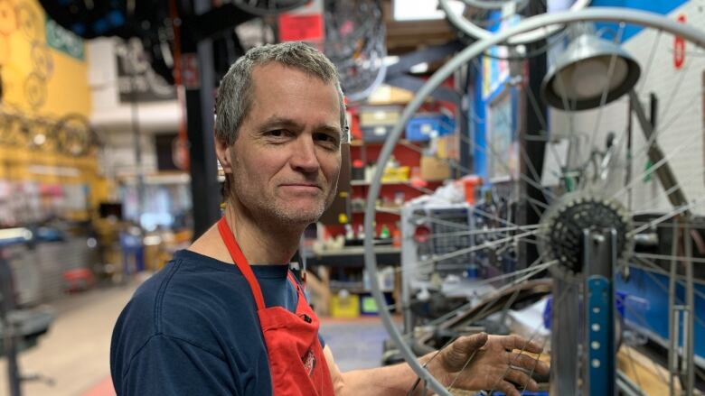 A man in a red apron stands next to a bike wheel in a shop.