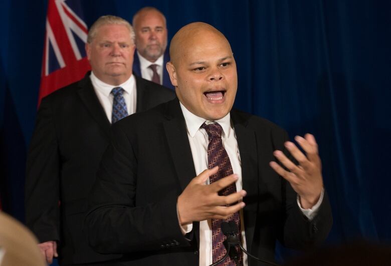 A man gestures while speaking at a microphone, as two men watch on from behind him.