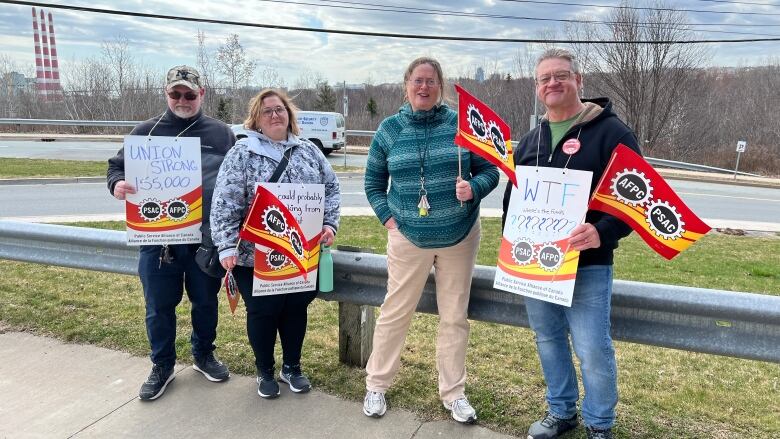 Four people holding signs stand against a fence.