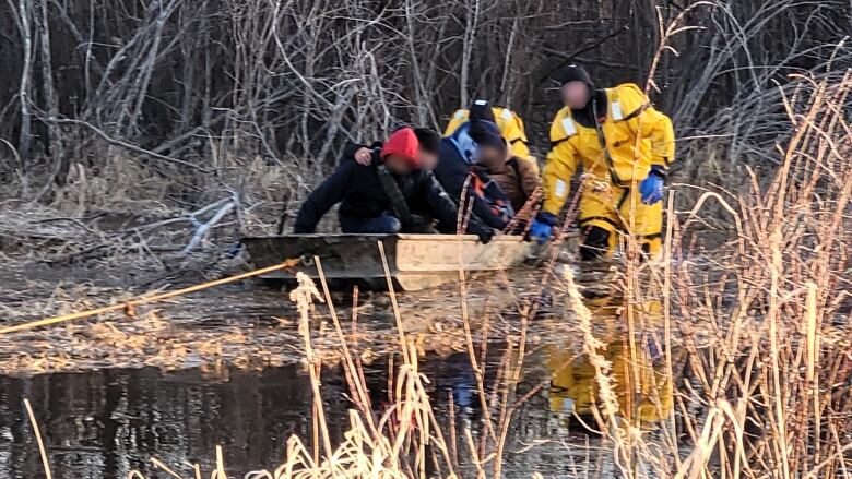 Rescuers in yellow drysuits help migrants get into a flat-bottomed boat in a wetland.