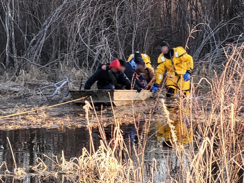 Rescuers in yellow drysuits help migrants get into a flat-bottomed boat in a wetland.