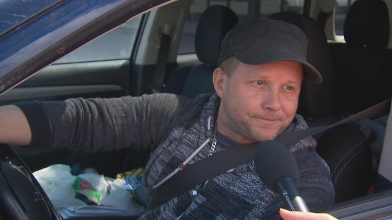 A man speaks into a microphone, seated in his car. 