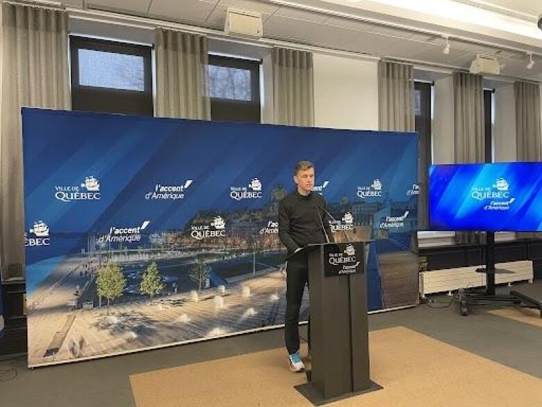 A man stands at a podium in front of a banner with the City of Quebec logo 