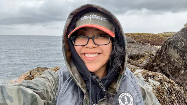 A woman with glasses is pictured wearing a hoodie in front of a sea and rocks.