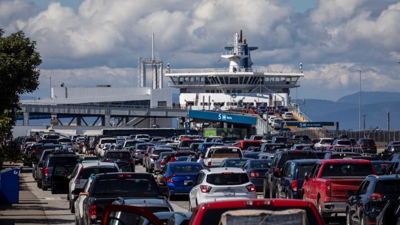 Hundreds of cars are waiting for a B.C. Ferry on a sunny day. The docked ferry is framed by the ocean and mountains.