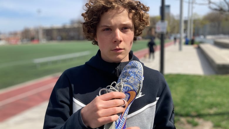 A teen boy holds a torn Nike soccer cleat next to a track surrounding a soccer pitch. 