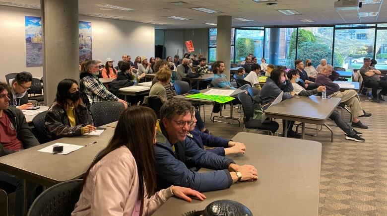 About 40 parents sit at tables watching a school board meeting, some holding protest signs.