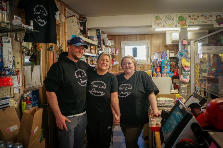 Three people in black shirts with the words Seal Cove Grocery on them smile at the camera behind the counter in a crowded convenience store.