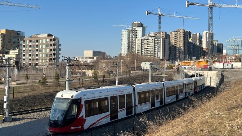 A red and white light rail train comes out of a tunnel with construction cranes and highrises in the background.