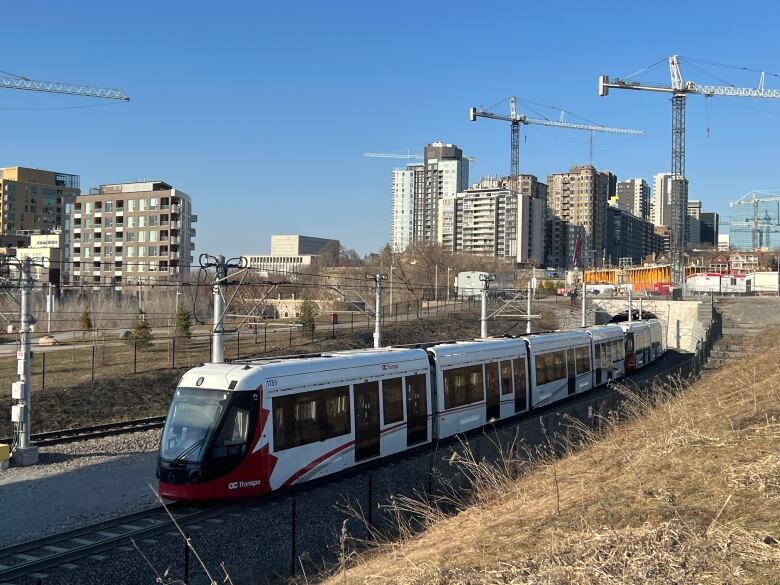 A red and white light rail train comes out of a tunnel with construction cranes and highrises in the background.