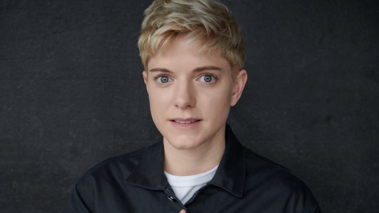 Head shot of comedian Mae Martin who's wearing a grey button-up shirt and standing against a plain grey wall.