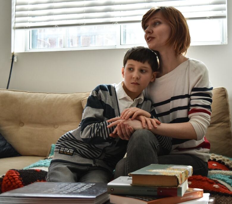 A woman and boy hold each other while sitting on a couch beside a table with books on it.