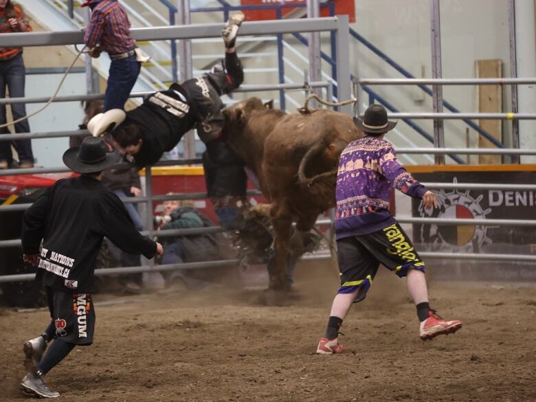 A bull rider flies through the air as two bullfighters approach in an attempt to distract it.