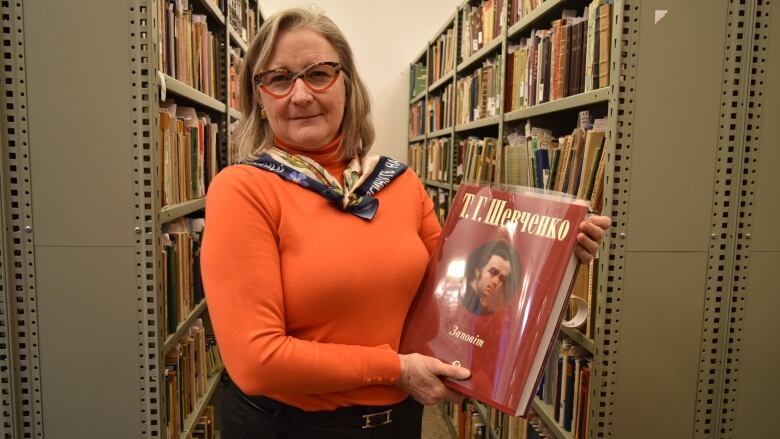 A woman holds a large book in front of bookshelves.