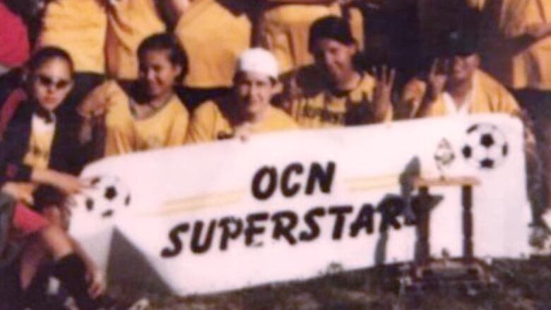 group of women on a soccer pitch they are wearing matching yellow uniforms, the women sat in the first row are holding up a banner that reads 