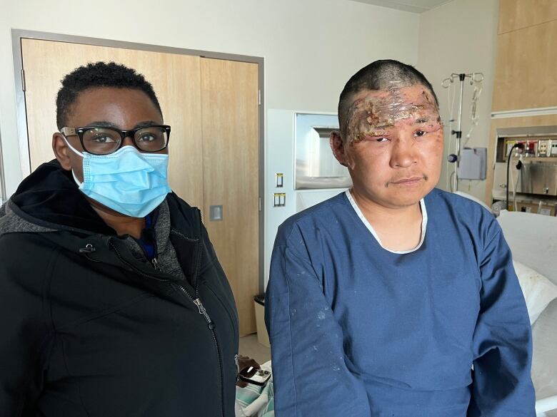 Woman in mask next to scarred man in hospital gown. 