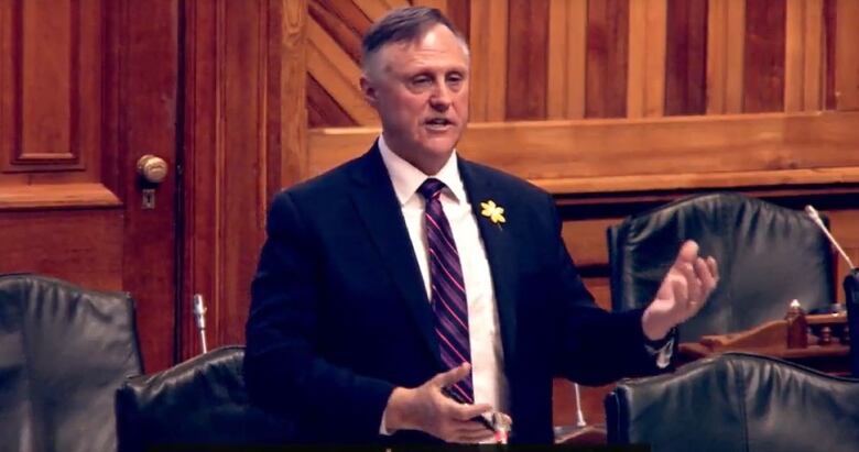 A man wearing a dark suit, white shirt and striped tie, holding a pen, speaking and gesturing with his hands.