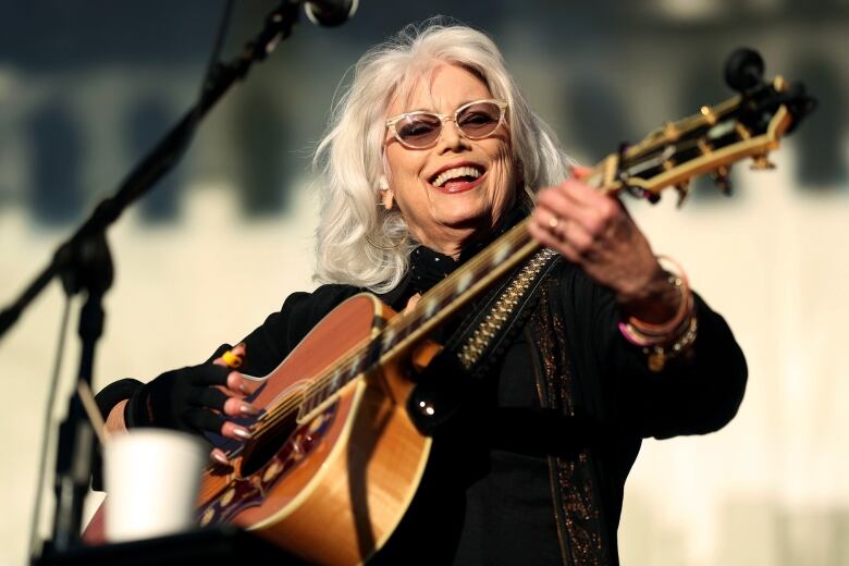 Wearing sunglasses and a dark shirt, a white-haired woman smiles while playing an acoustic guitar.