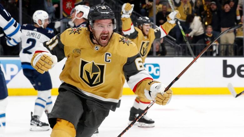 A male ice hockey player pumps his right fist while shouting in celebration as a teammate raises his arms into the air behind him.