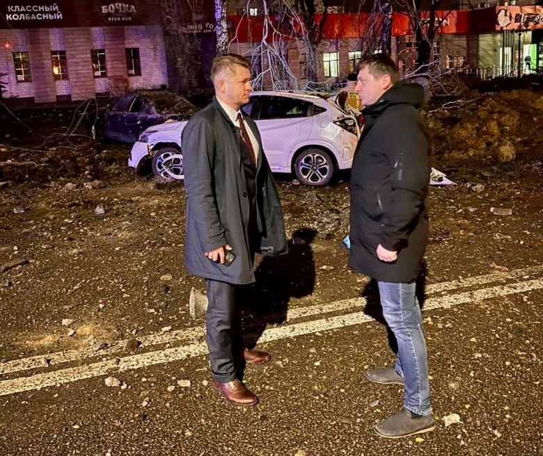 Two men are shown in conversation on a street in a nighttime photo, with damage and debris seen in the background.