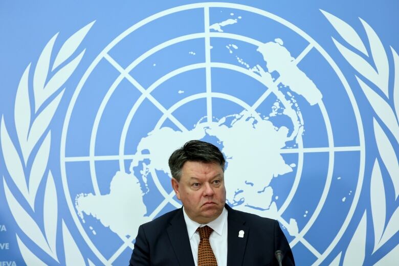 A man frowns in front of a blue and white logo featuring the world as seen in a flattened projection from above the north pole.