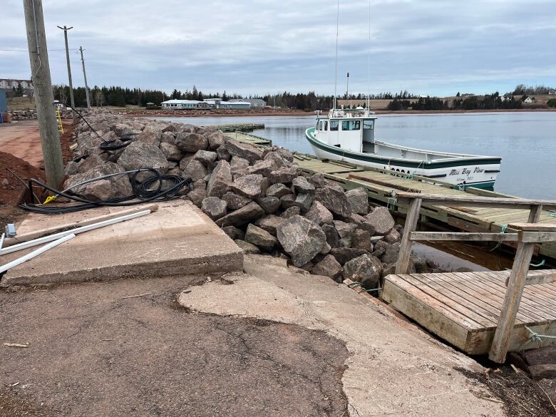 A pile of rocks line the shoreline with a fishing boat tied up in the harbour 