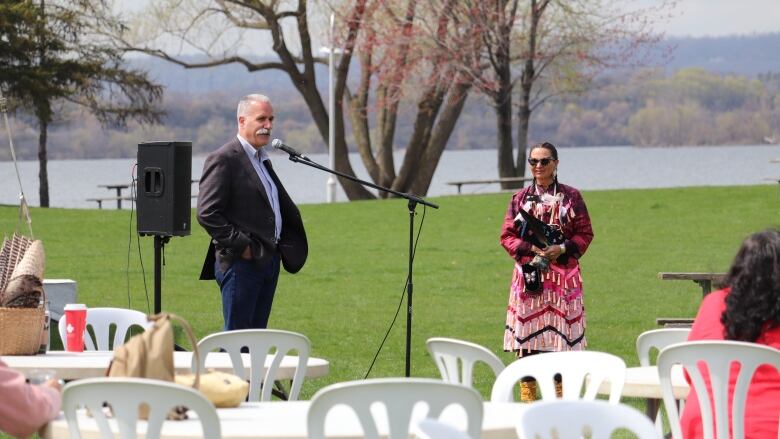 A man speaking into a microphone while facing a woman in an Indigenous jingle dress. 