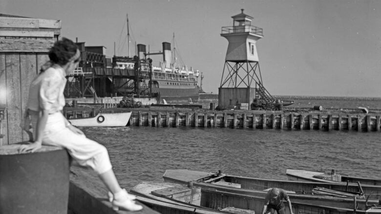 woman sitting on pier in black and white photo from 1936 looking out at ferry, dock and lighthouse